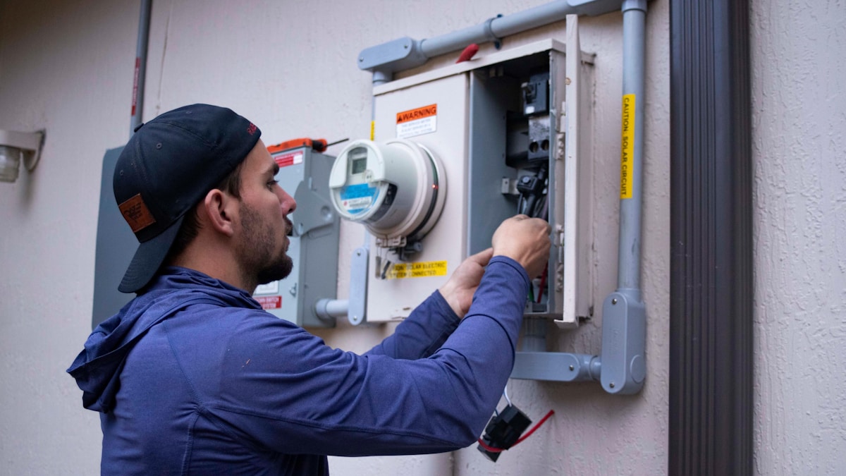 Un technicien en uniforme bleu vérifie un compteur électrique mural.