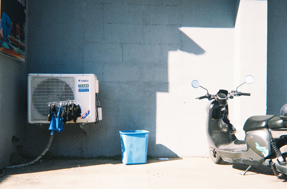 Unité de climatisation, scooter, poubelle bleue, mur gris, ombre lumineuse.