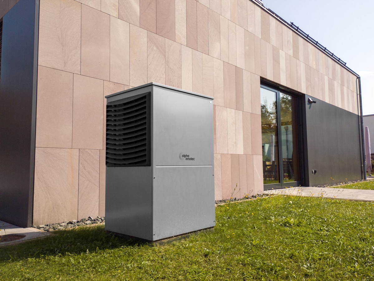 Unité de climatisation extérieure Alpha Innotec sur gazon devant un bâtiment moderne.