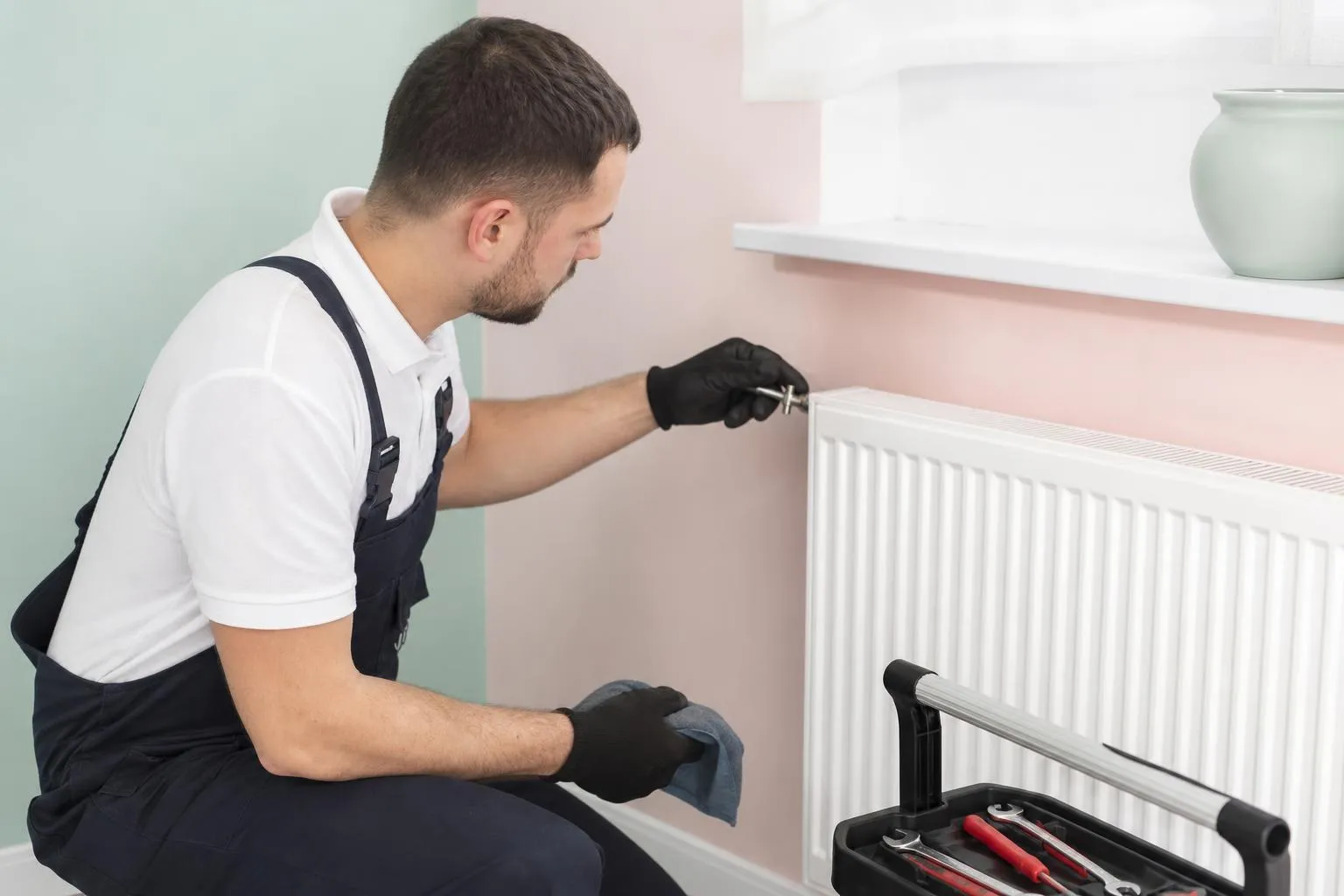 Un homme en uniforme de travail répare un radiateur à l'intérieur d'une pièce.